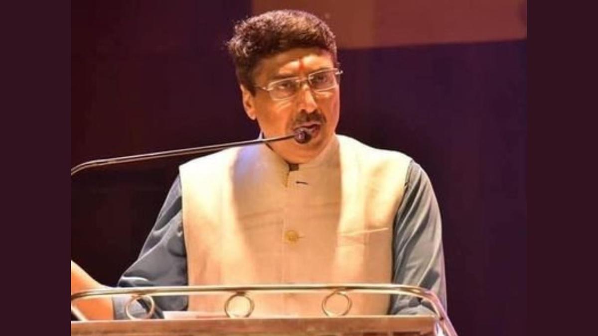 Deepak Mhaskey speaking into a microphone from a lectern at a public event, wearing a light Nehru jacket.