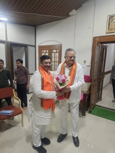 Deepak Mhaskey in a checked shirt presenting a bouquet of pink and cream flowers to a leader in a brown Nehru jacket, in front of a large black-and-white photograph of a crowd.