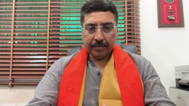 Deepak Mhaskey seated at a desk in an office, wearing a grey jacket with a saffron and yellow stole, speaking to camera in front of window blinds.